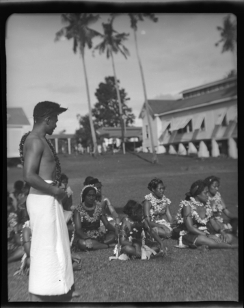 Group of women dressed alike sitting on grass, and man in white skirt standing