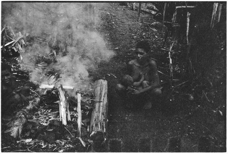 Woman tends a fire, while nursing her infant