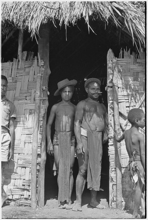 Men stand in doorway of government rest house in Tsembaga, one wears western-style hat and another wears luluai badge