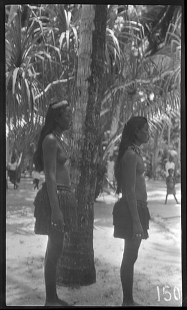 Two women in grass skirts and necklaces, rings, and head piece