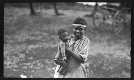 Woman holding infant, carriage in background