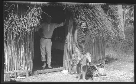 Man in doorway of a Cook Islands house, with woman and food baskets outside