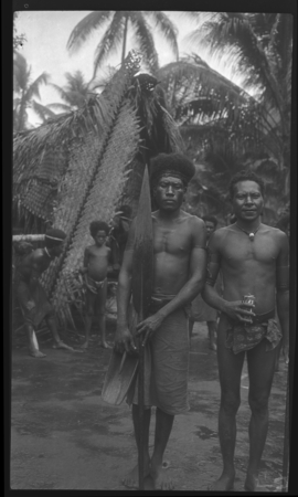 Two Trobriand men in front of a house; man on left is holding a canoe paddle, and a water bailer