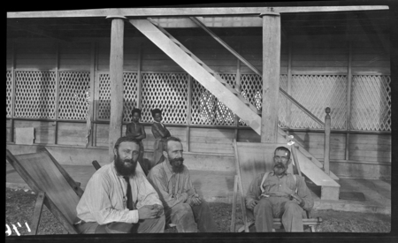 Catholic priests sitting near a mission building, young Papuan men in background, Yule Island