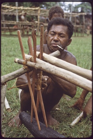 Canoe-building: Mogiovyeka attaches outrigger float to a canoe, lashing with lengths of vine, he is smoking