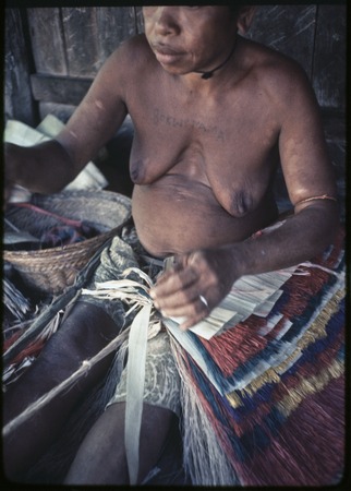Weaving: woman makes skirt out of banana leaf fibers, some dyed red, blue or yellow, woman has word tattooed on her chest
