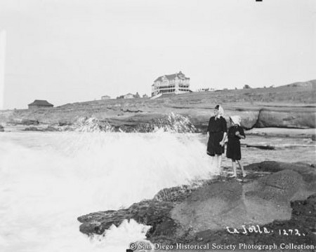 Two girls in bathing suits standing on rock at La Jolla beach, La Jolla Park Hotel in background