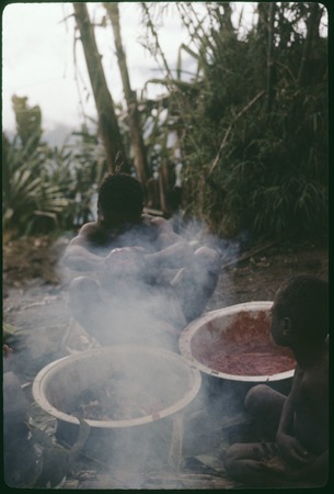 Food preparation: man squeezes red pandanus fruits, producing red juice seen in bowl (r)