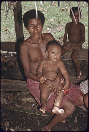 Woman holding an infant, smiling boy in background, woman&#39;s hand is tattooed