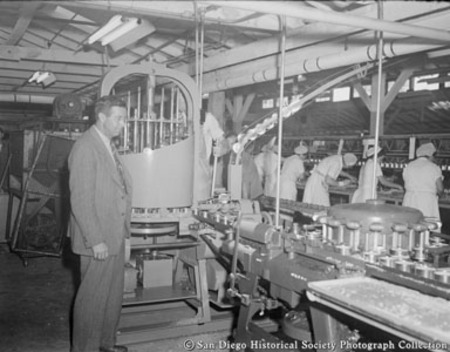 Interior view of San Diego cannery showing workers and man in suit standing next to fish filling machine manufactured by S...