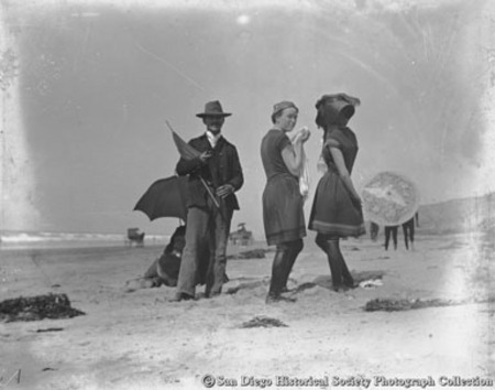 Two young women in bathing suits standing with man holding parasol on beach at Coronado&#39;s Tent City