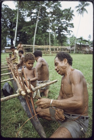 Canoe-building: men use lengths of vine to attach an outrigger float to a canoe, young child observes