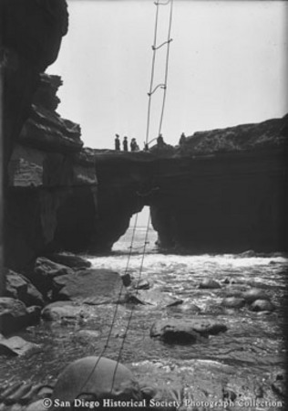 Rope ladder hanging from cliff to shore, with people standing on natural bridge rock formation in background, Sunset Cliffs