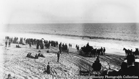 Crowd of people and horse drawn carriages on Coronado beach, two people in ocean surf