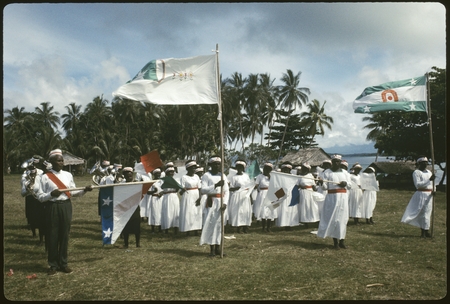 Group, formal: people in white dress with headbands and red sashes, assortment of flags, and musicians to their right