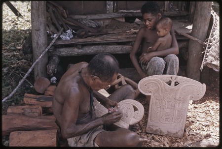 Carving: M&#39;lapokala creates a design on canoe prowboard, next to carved splashboard, boy and infant sit nearby watching