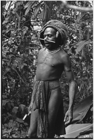 Man wearing barkcloth cap, netted loincloth, beaded necklace, woven armband and belt, carries an axe