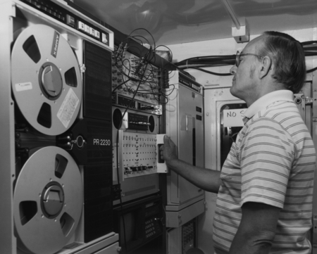 Technician stands in front of the state of the art tape recorders and electronic equipment used in recording and processin...