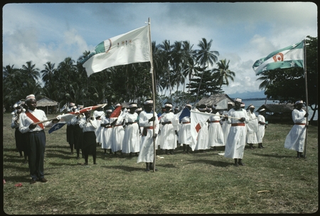 Group, formal: people in white dress with headbands and red sashes, assortment of flags, and musicians to their right