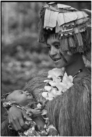 New mother, holding infant, wears long fiber shawl, flower garland, and skirt valuable on head, paint on face