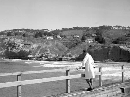A unidentified person standing on the Scripps pier enjoying a view of the ocean and the coastline. Note the cottages and t...