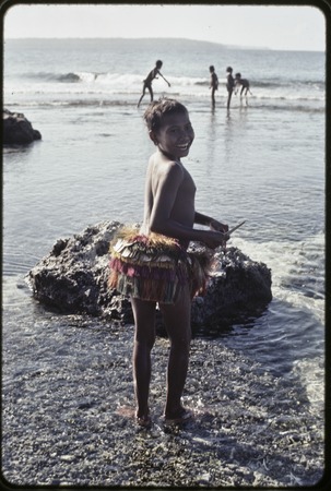 Girl in short fiber skirt walks on the fringing reef near Wawela village at low tide, other children fish on edge of coral...