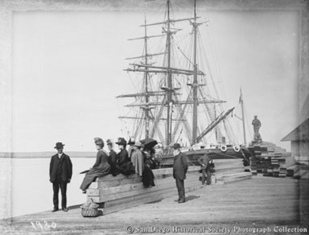 View of Santa Fe Wharf showing women sitting on stack of lumber and docked sailing ship