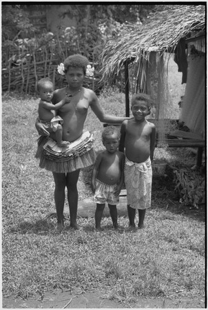 Adolescent girl wearing short fiber skirt and necklace, holds infant, younger children stand nearby