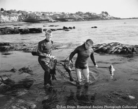 Members of La Jolla Skinsters Diving Club Michael Carnohan and Frank Snodgrass standing in surf with spear fishing catch o...