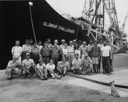 Scientists and technicians of Deep Sea Drilling Project, Leg unknown, on a dock with the D/V Glomar Challenger (ship) whil...