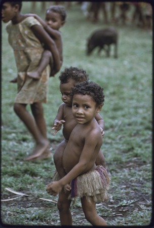 Children carry babies and younger children, girl in foreground wears short fiber skirt