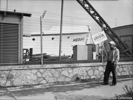 Labor union member with picket sign in front of boat and ship building company, boat Heroic in background