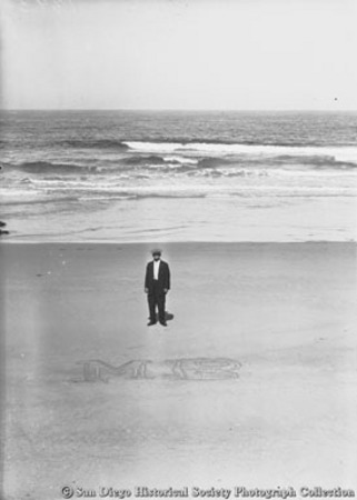 Man standing on beach behind letters &quot;MB&quot; written in the sand