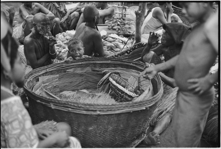 Mortuary ceremony, Omarakana: large basket of exchange valuables, banana leaf bundles stacked, woman (l) smokes