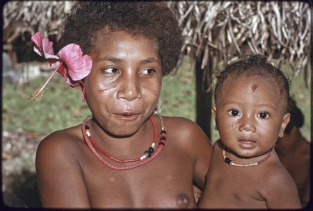 Adolescent girl, hibiscus flower in hair and dabs of betel nut paste on cheeks, holds an infant, both wear shell necklaces