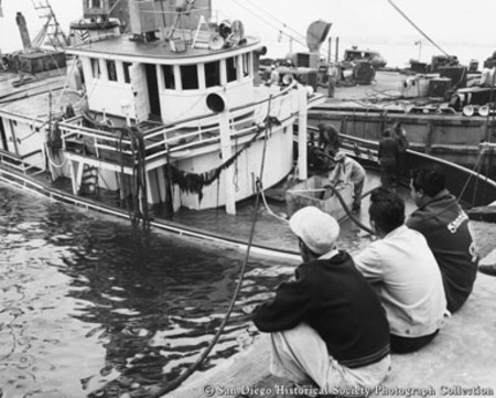 Three men sitting on dock watching salvaging operations for sunken tuna boat Normandie