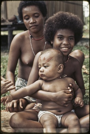 Adolescent girl holding an infant with betel nut paste dabbed on face, woman sits behind, all wear shell necklaces