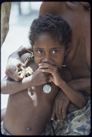 Young child wearing a coin necklace gazes at camera, stands near a woman