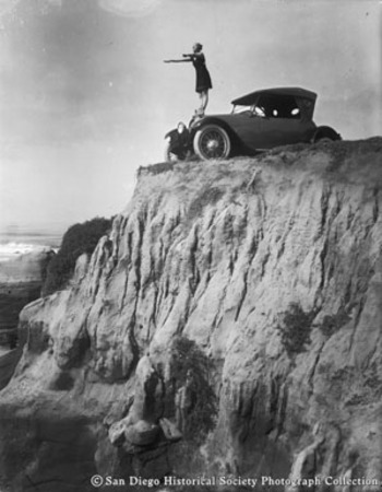 Woman in bathing suit pretending to dive off automobile parked on cliff overlooking Pacific Ocean