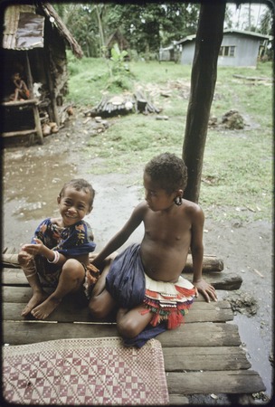 Children sitting on house veranda next to woven mat, girl (r) wears short fiber skirt and turtle-shell earrings,