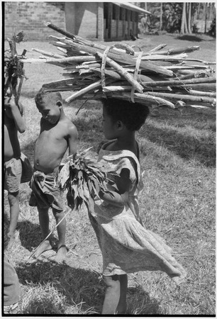 Children, girl (r) balances load of firewood on her head and carries bundle of greens, church in background