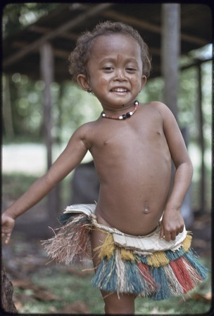 Young girl, Imala (daughter of Mogiovyeka), wearing short fiber skirt and shell necklace, dabs of betel-nut paste on her c...