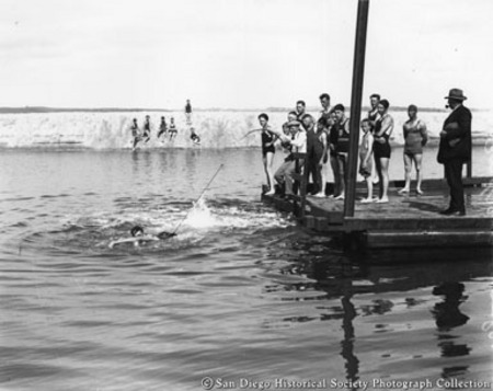 [Humorous photograph of men and boys on diving platform watching man with fishing rod trying to reel in swimmer]