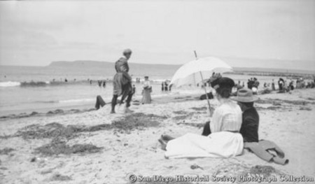 Beach scene at Coronado&#39;s Tent City showing woman holding parasol and man sitting together in sand