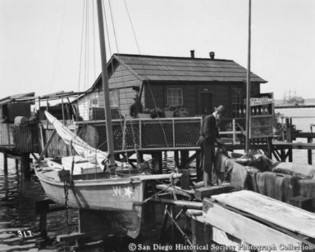 View of San Diego waterfront showing house, man with two boats, and sign reading &quot;Saw filing and shell grinding&quot;