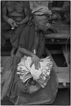 Mortuary ceremony: mourning woman with shaved head, holds with banana leaf bundles (wealth items)