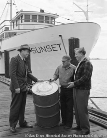 Three men standing around oil drum, docked tuna boat Sunset in background