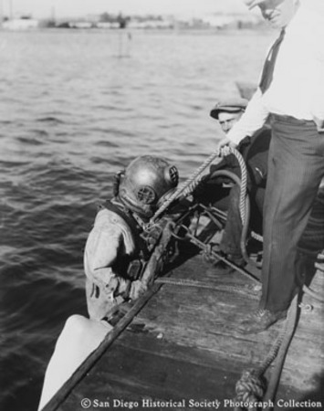 Two men on dock lowering woman in diving suit into San Diego Bay