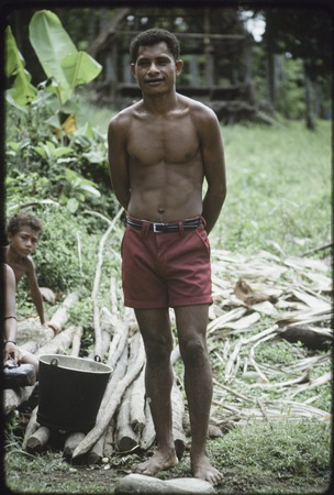 Mowaroka stands next to bucket and poles from which bark has been removed, bark (background) will be made into tapa