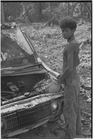 Truck with damaged front end, adolescent boy stands beside engine with improvised repairs